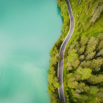 Aerial view of a winding road beside a green forest and turquoise lake