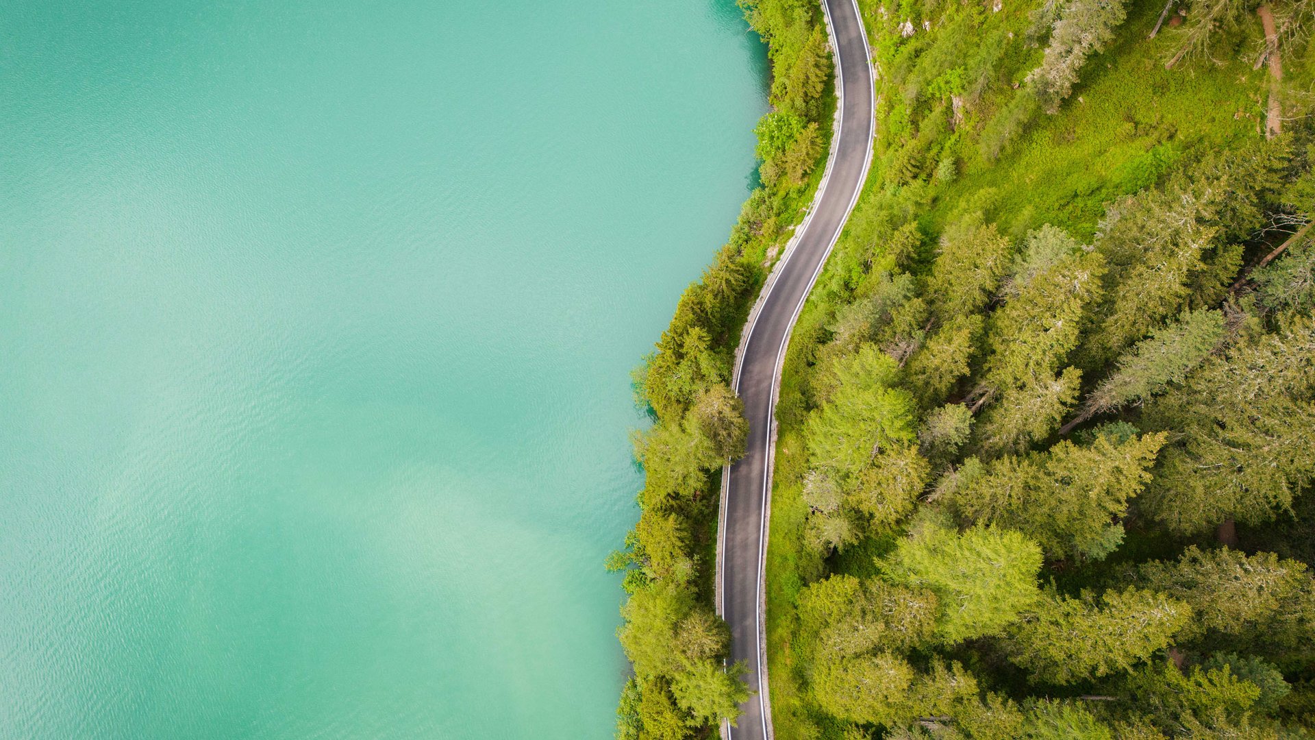Aerial view of a winding road beside a green forest and turquoise lake
