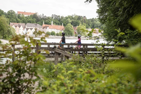 Entdeckungsreise Oberbayern © Velontour Zwei Radfahrer auf einem Holzsteg mit Blick auf einen Fluss und Häuser