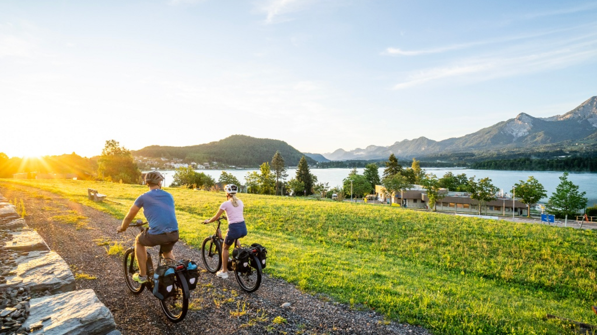 Two cyclists on a path beside lake and mountains at sunset