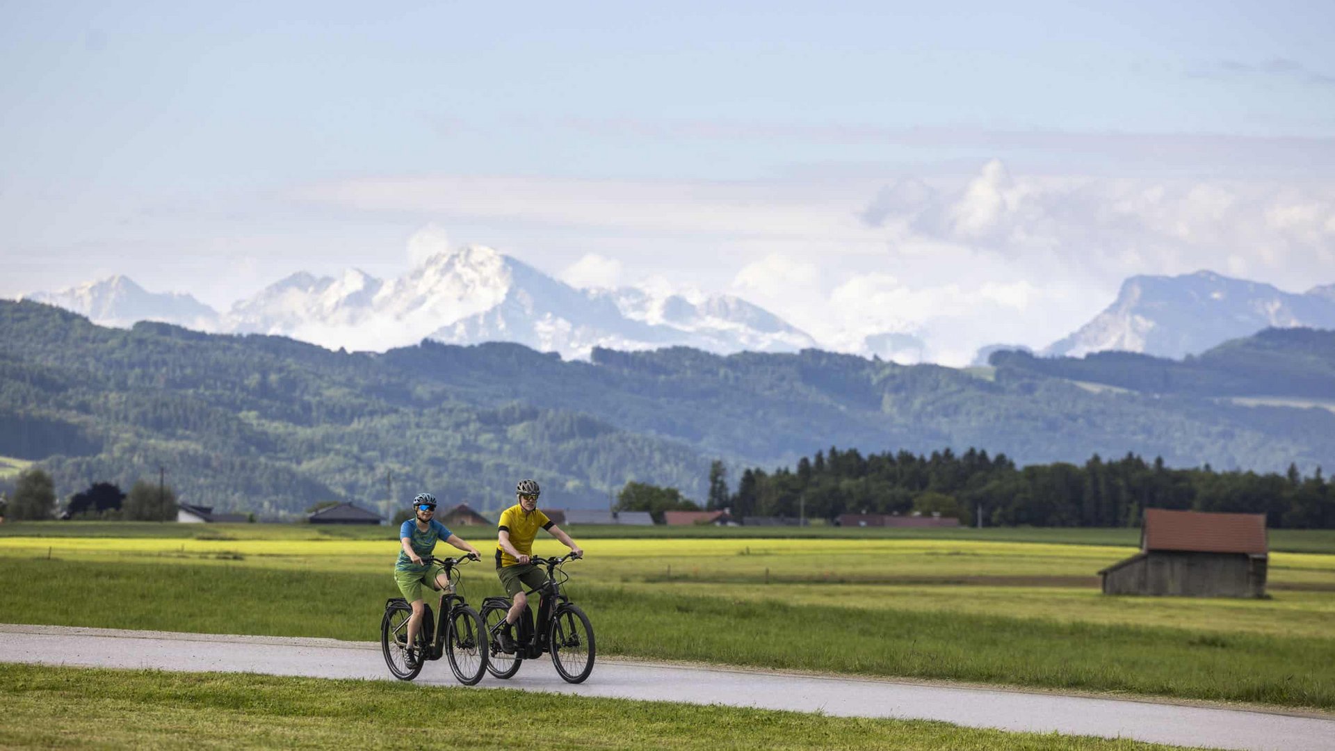 Two cyclists riding on a path through green fields with mountains in the background
