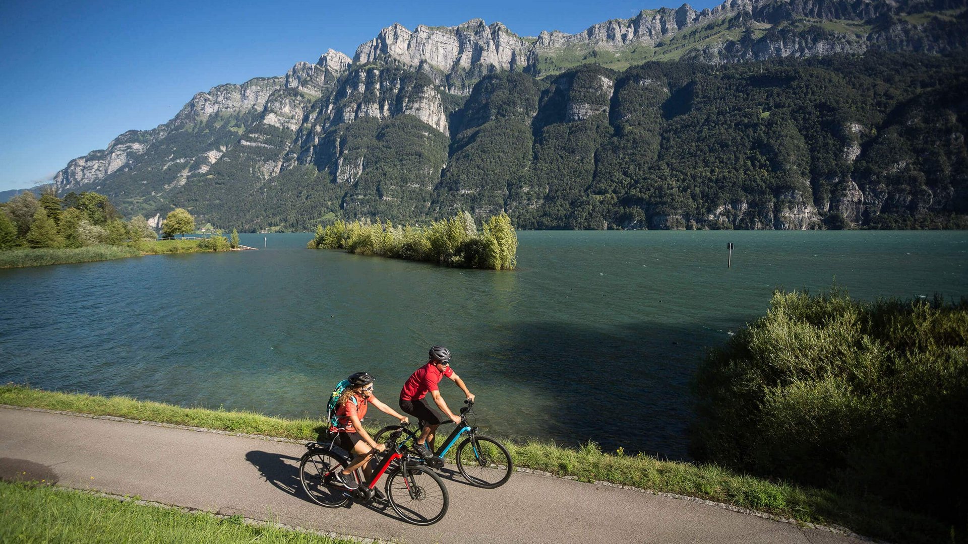 Zwei Radfahrer fahren auf einem Weg neben einem See und Bergen im Hintergrund
