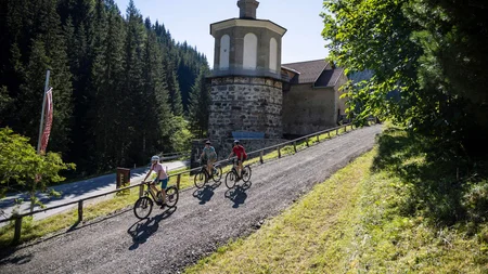 Drei Radfahrer auf einem Weg bergauf neben historischem Turm im Wald