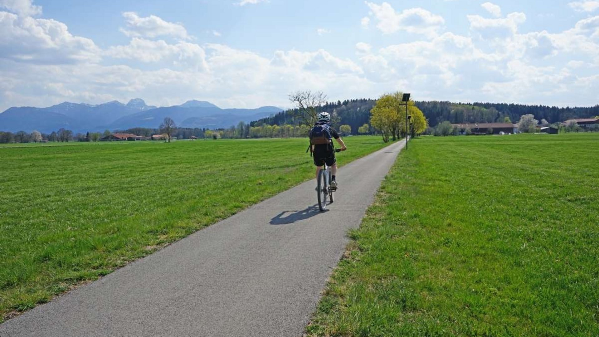 Person cycling on a path through green fields with mountains in the background