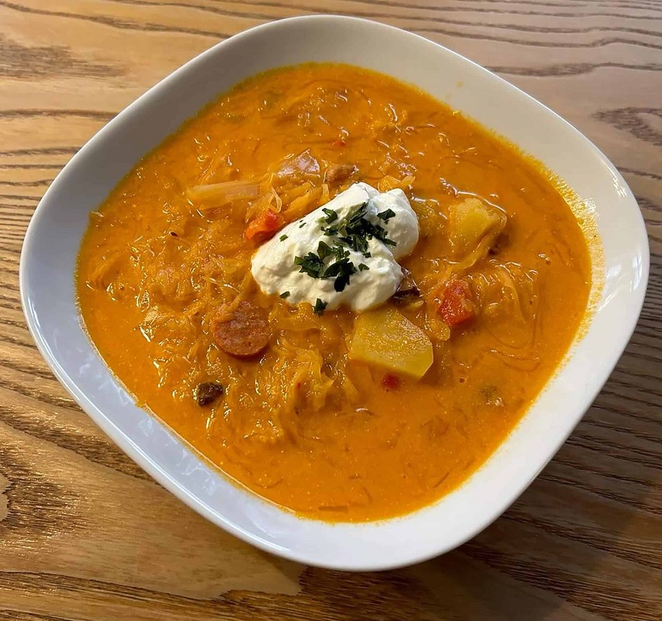 Bowl of sauerkraut soup with potatoes, sausage, and sour cream on wooden surface.