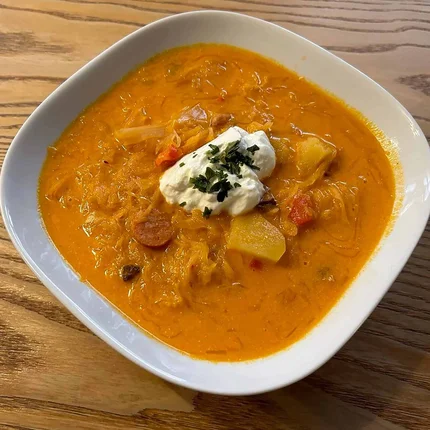 Schüssel mit Sauerkrautsuppe, Kartoffeln, Wurst und saurer Sahne auf Holzuntergrund.
