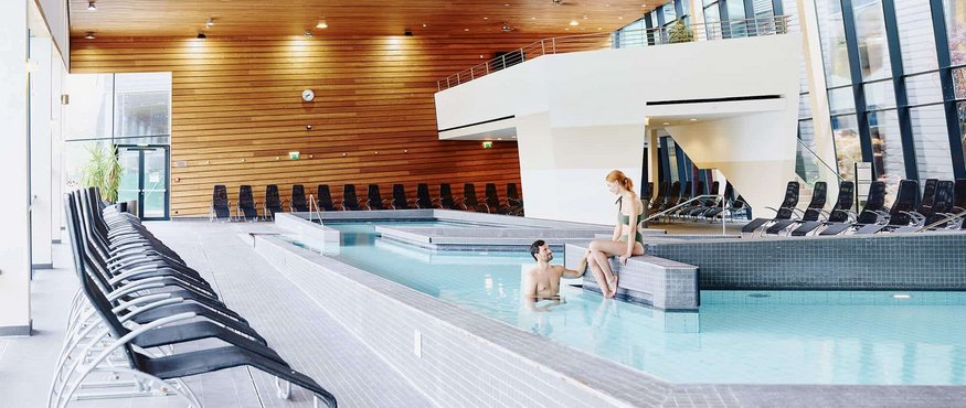 Interior of a modern indoor pool with people in water and brown wooden ceiling.