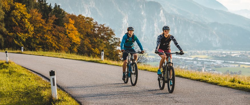 Two cyclists riding on mountain road with autumn trees and mountains behind