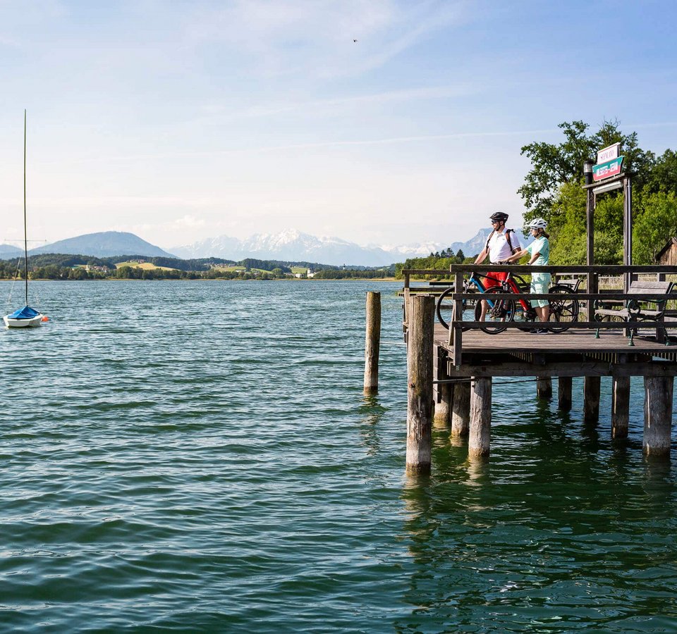 Zwei Radfahrer auf einem Steg am See mit Bergpanorama bei klarem Himmel