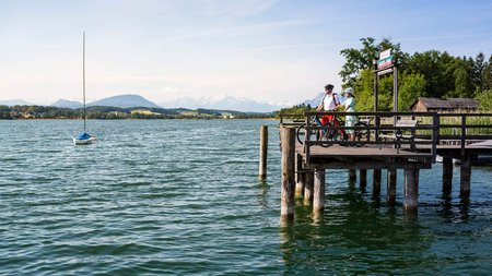 Zwei Radfahrer auf einem Steg am See mit Bergpanorama bei klarem Himmel