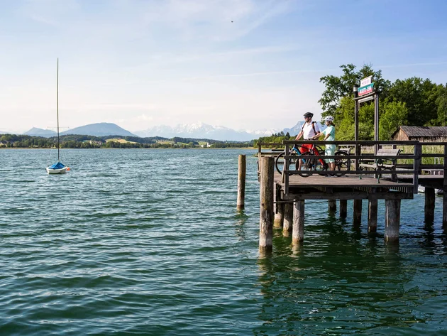 Zwei Radfahrer auf einem Steg am See mit Bergpanorama bei klarem Himmel