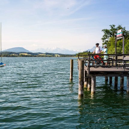 Zwei Radfahrer auf einem Steg am See mit Bergpanorama bei klarem Himmel