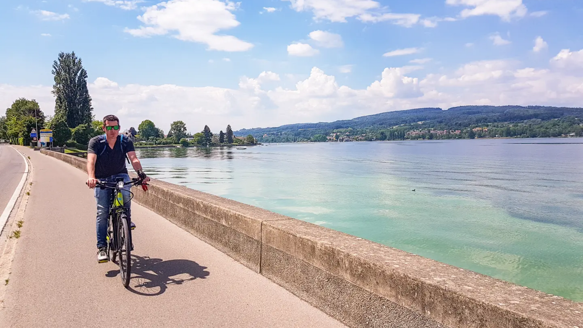 Man cycling on path beside lake with hills in background