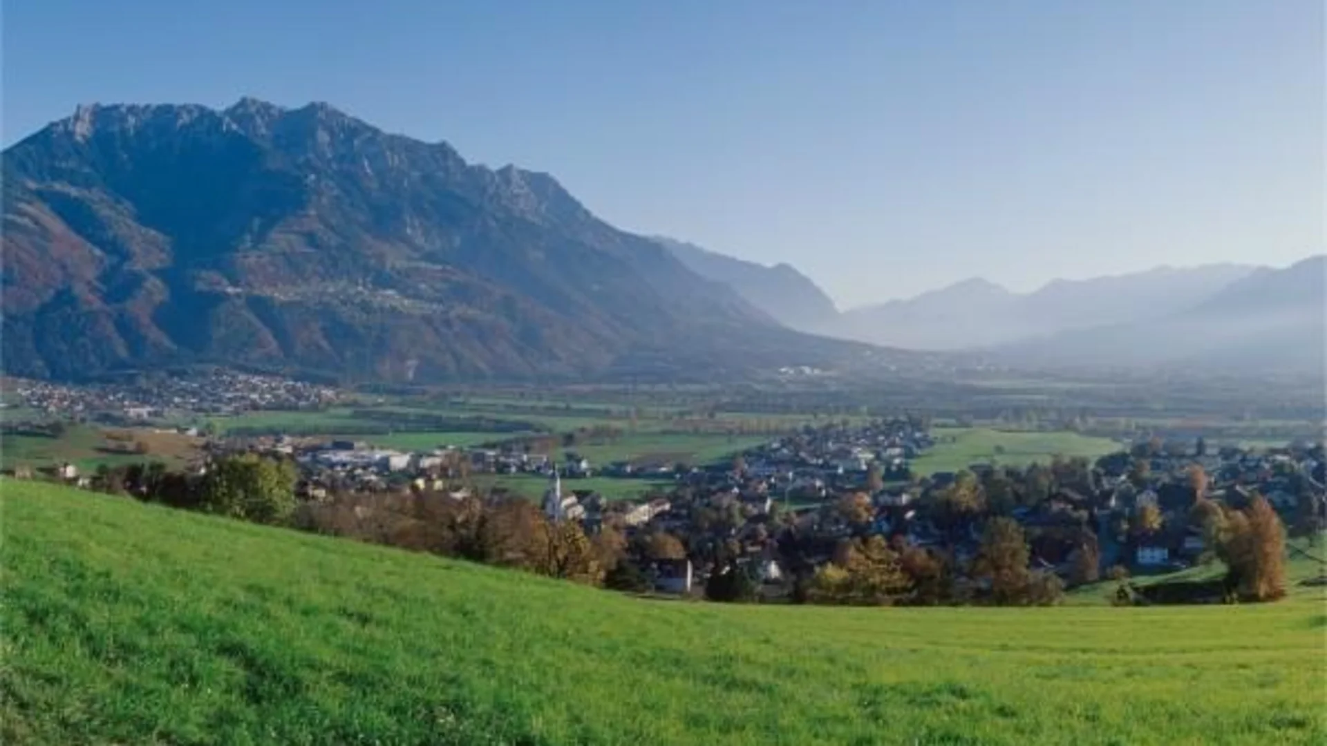 Blick auf eine Stadt im Tal mit grünen Wiesen und Bergen im Hintergrund