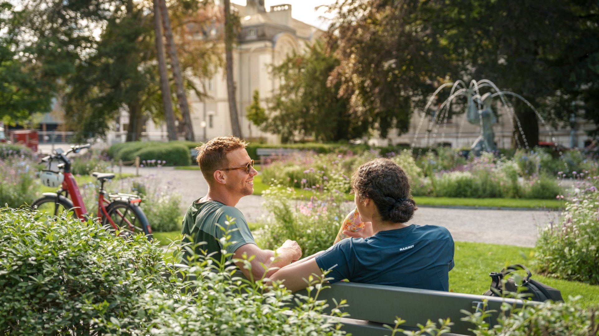 Two people sitting on a park bench next to a bicycle on a sunny day