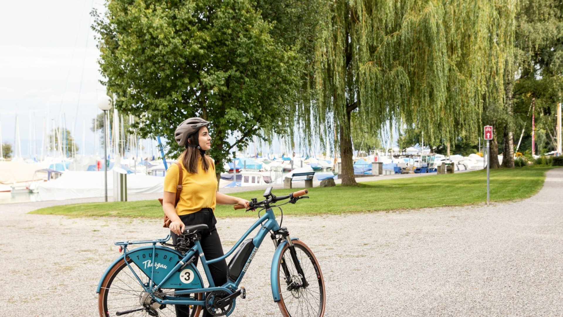 Woman with bicycle on a park path with trees and boats in the background