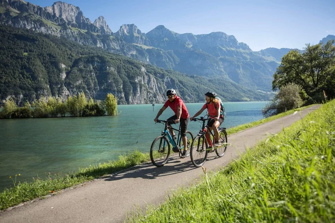 Zwei Radfahrer auf einem Weg neben einem See mit Bergen im Hintergrund