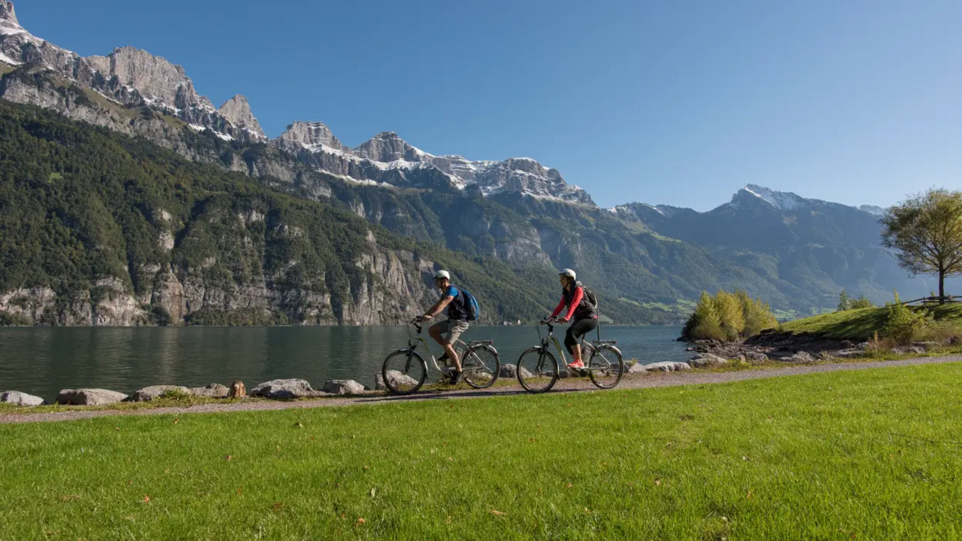 Two cyclists by a lake with snow-capped mountains under clear sky