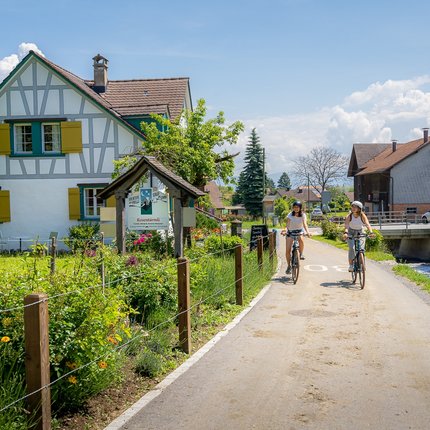 Two people cycling on a path next to a stream and a half-timbered house.
