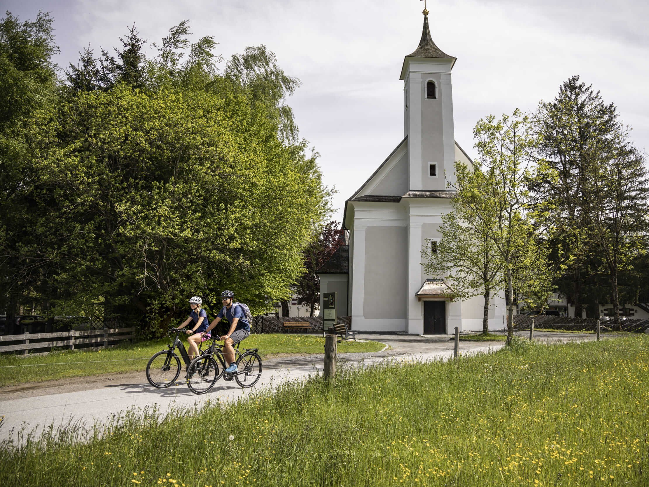 Zwei Radfahrer fahren auf einem Weg neben einer kleinen Kirche