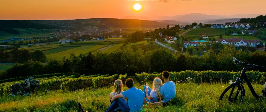 Vier mensen zitten in gras en kijken naar zonsondergang over dorp