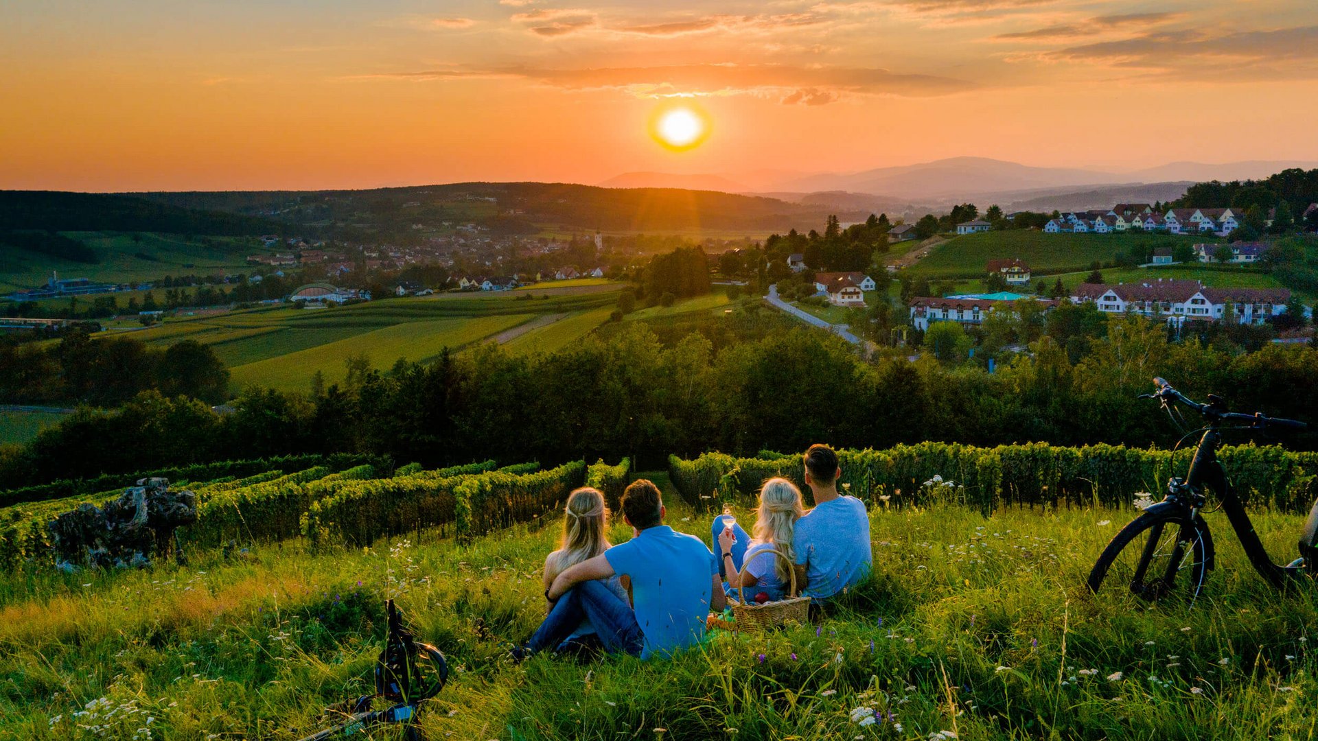 Four people sitting on grass watching sunset over village and hills