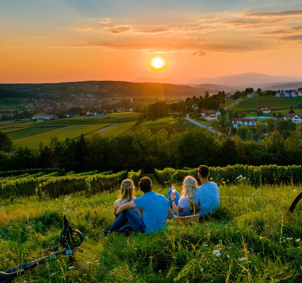 Genuss mit jedem Tritt im Thermen- & Vulkanland © TV Thermen & Vulkanland - Mias Photoart Vier Personen sitzen auf einer Wiese und schauen auf den Sonnenuntergang über dem Dorf