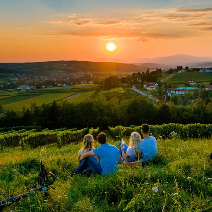 Genuss mit jedem Tritt im Thermen- & Vulkanland © TV Thermen & Vulkanland - Mias Photoart Vier Personen sitzen auf einer Wiese und schauen auf den Sonnenuntergang über dem Dorf