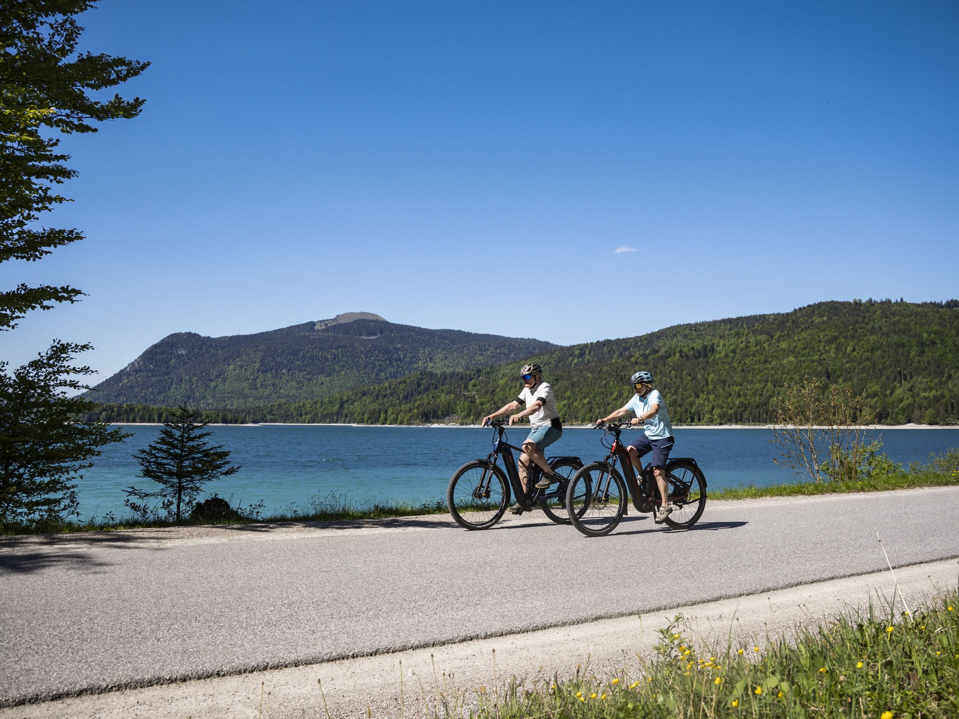 Two cyclists riding by a lake with mountains in the background