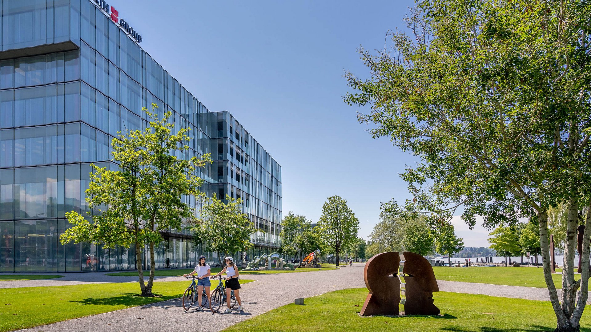 Modern glass building and sculpture in park with two cyclists on sunny day