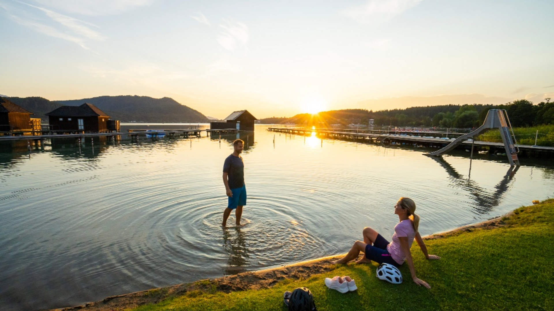 People by lake shore at sunset with slide and huts in background