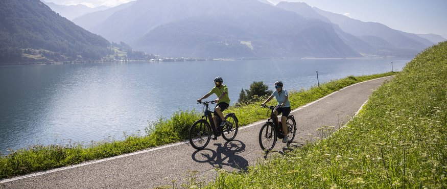Two cyclists on lakeside path with mountains behind
