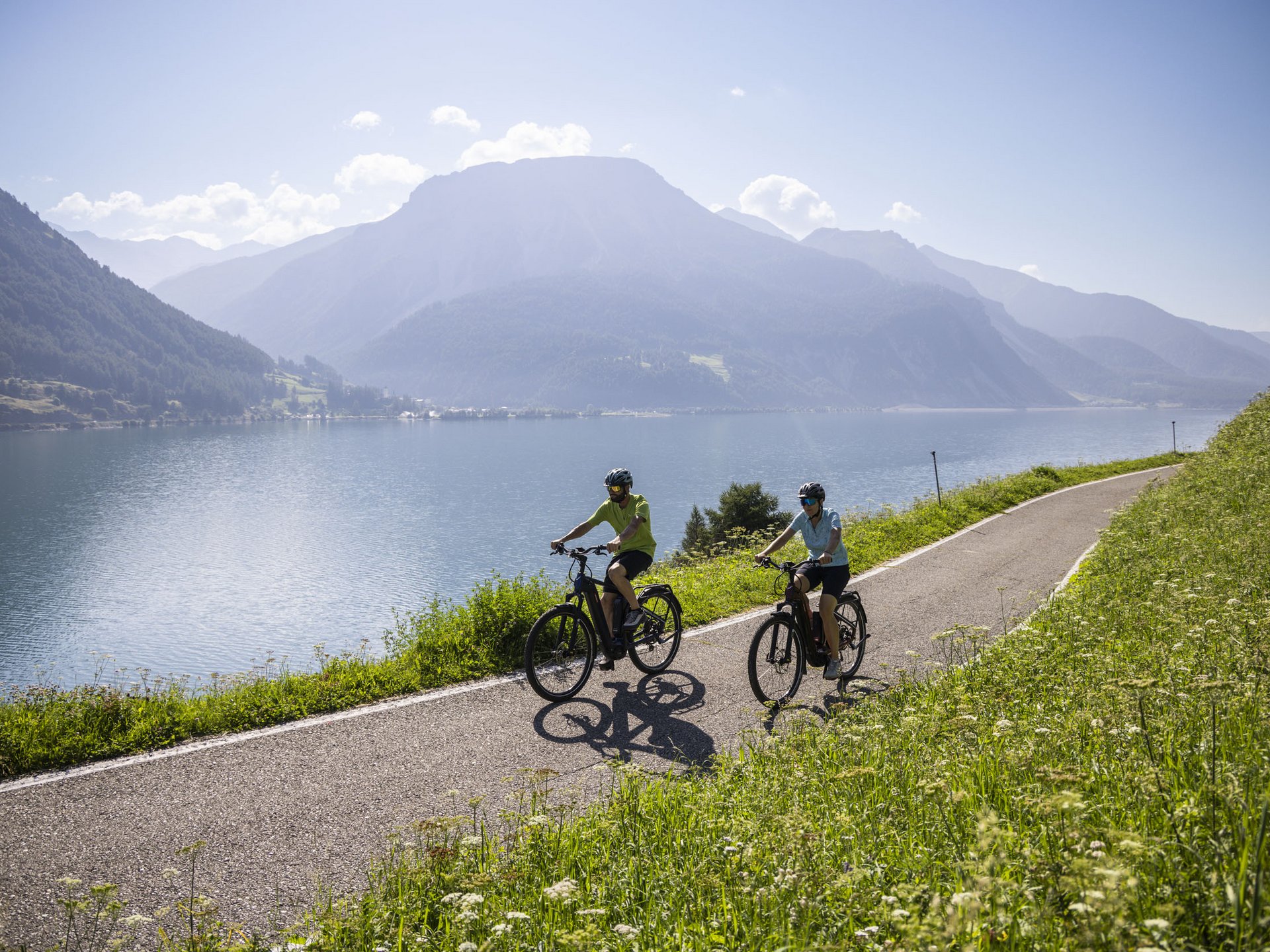 Two cyclists on lakeside path with mountains behind
