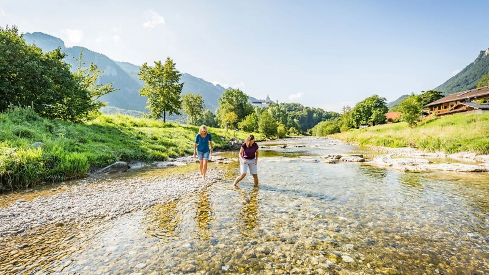 Twee vrouwen lopen blootsvoets door een rivier omringd door bergen en groen