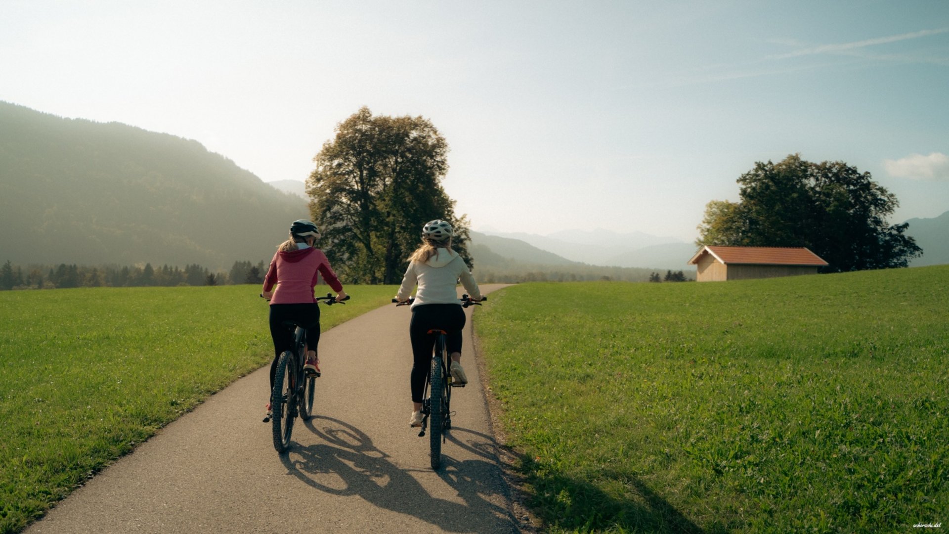 Two women cycling on a path through green landscape with mountains behind