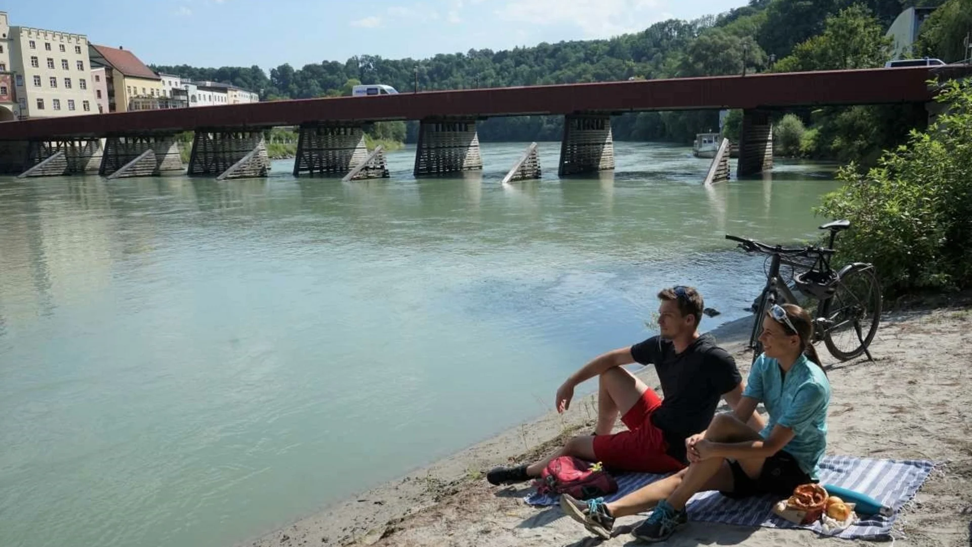 Paar sitzt am Flussufer mit Brücke und Fahrrad im Hintergrund