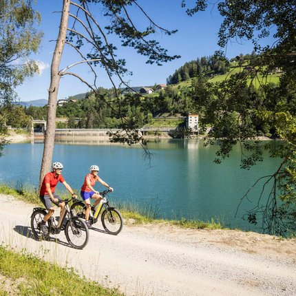 Zwei Radfahrer auf einem Weg neben einem See und Hügeln bei Sonnenschein