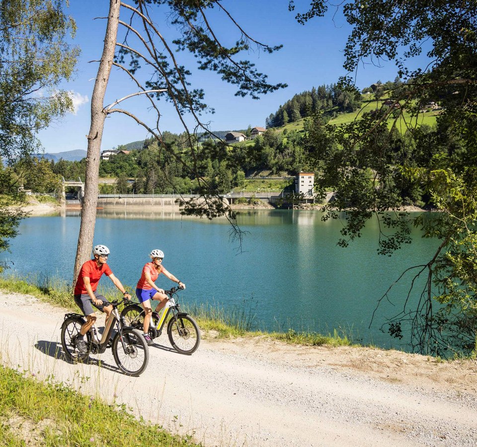 Zwei Radfahrer auf einem Weg neben einem See und Hügeln bei Sonnenschein