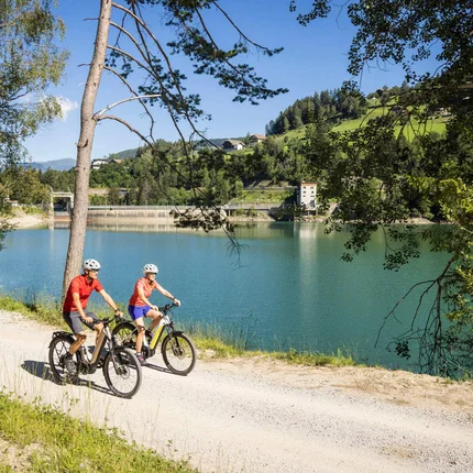 Zwei Radfahrer auf einem Weg neben einem See und Hügeln bei Sonnenschein