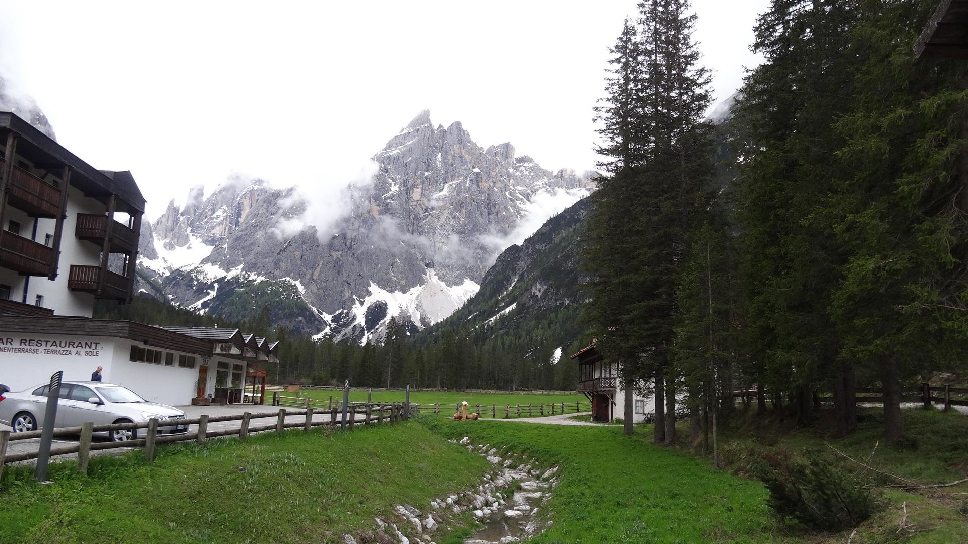 Mountain landscape with snowy peaks, forest edge, and buildings near a stream