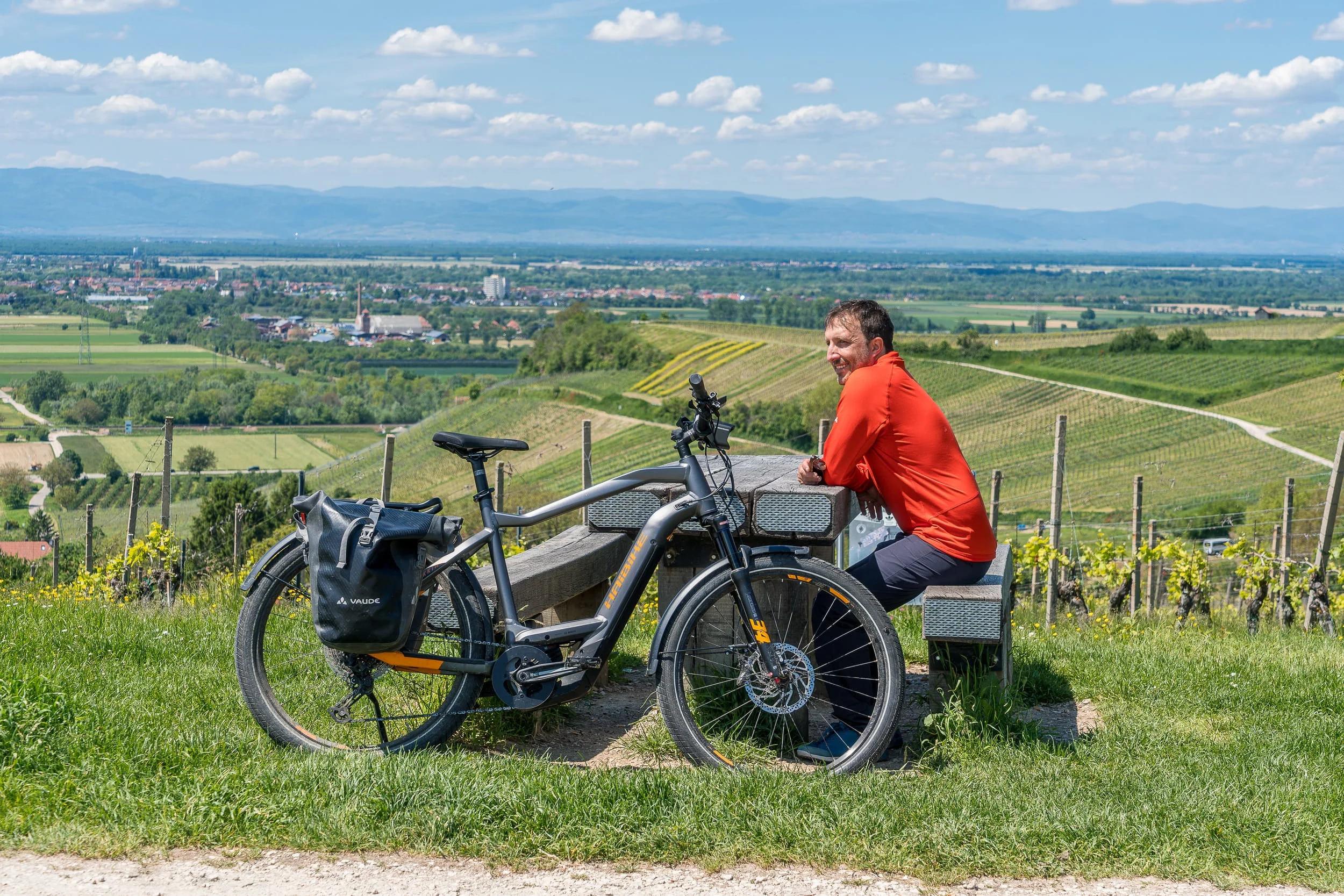 Mann in rotem Hemd sitzt auf Bank neben Fahrrad mit Aussicht auf Weinberge