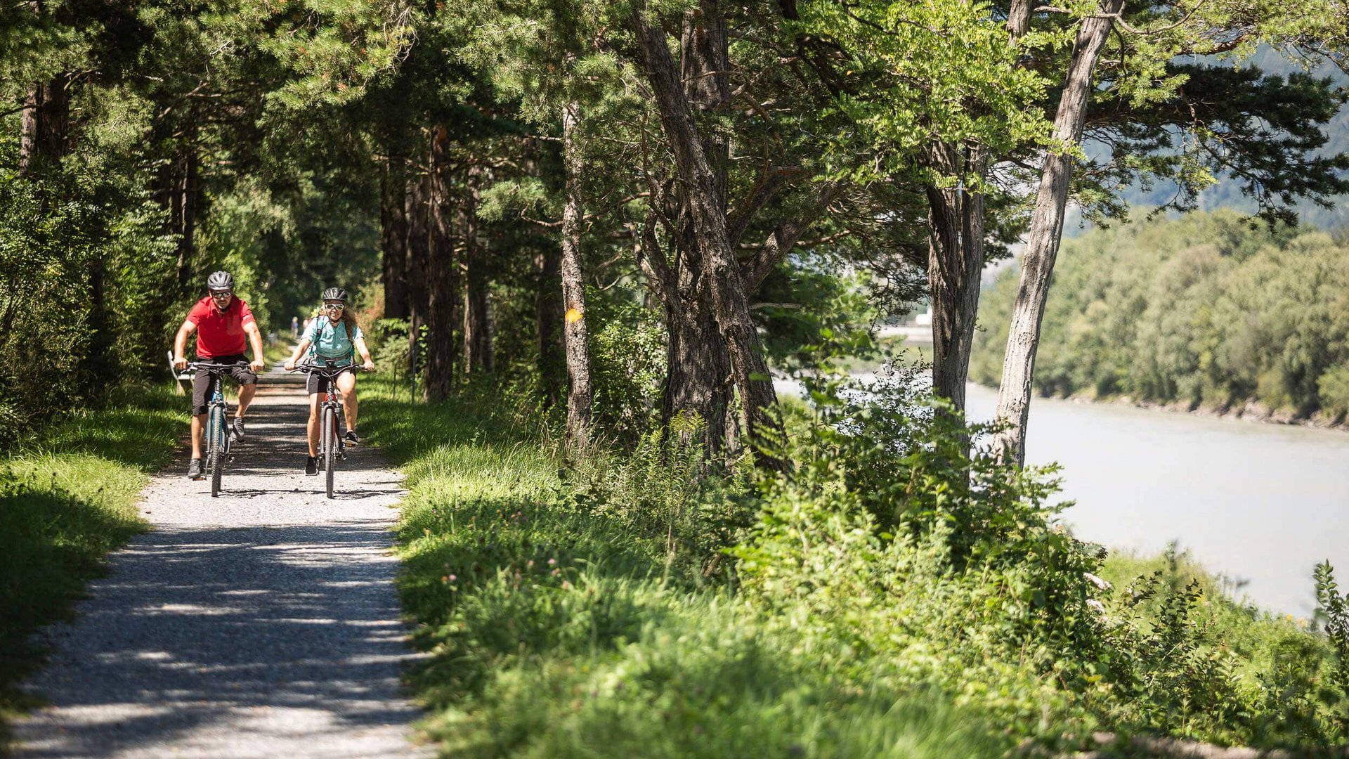 Two cyclists riding on a shaded forest path beside a river.