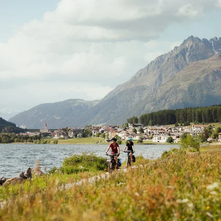 Twee fietsers langs een meer met bergen en dorp op de achtergrond