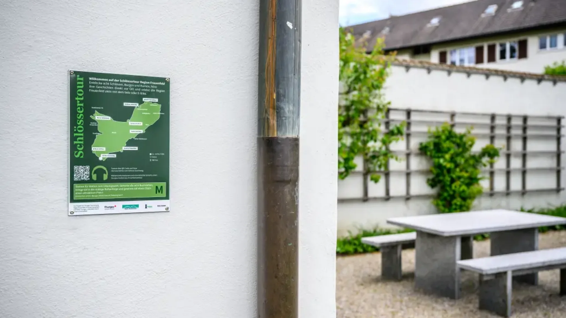 Tour-Detail © Balz Kubli Infotafel Schlosssertour an einer weißen Wand neben Außenbereich mit Tisch und Bank