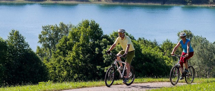 Tannberg Radrunde © Agentur Dreirad Zwei Radfahrer fahren auf einem Weg neben einem See und Bäumen