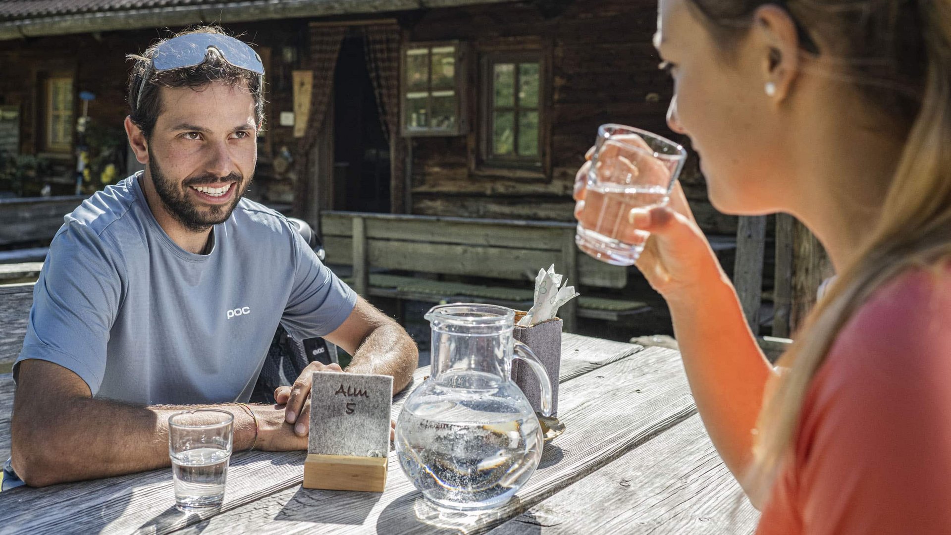 Two people drinking water at a wooden table outside a cabin