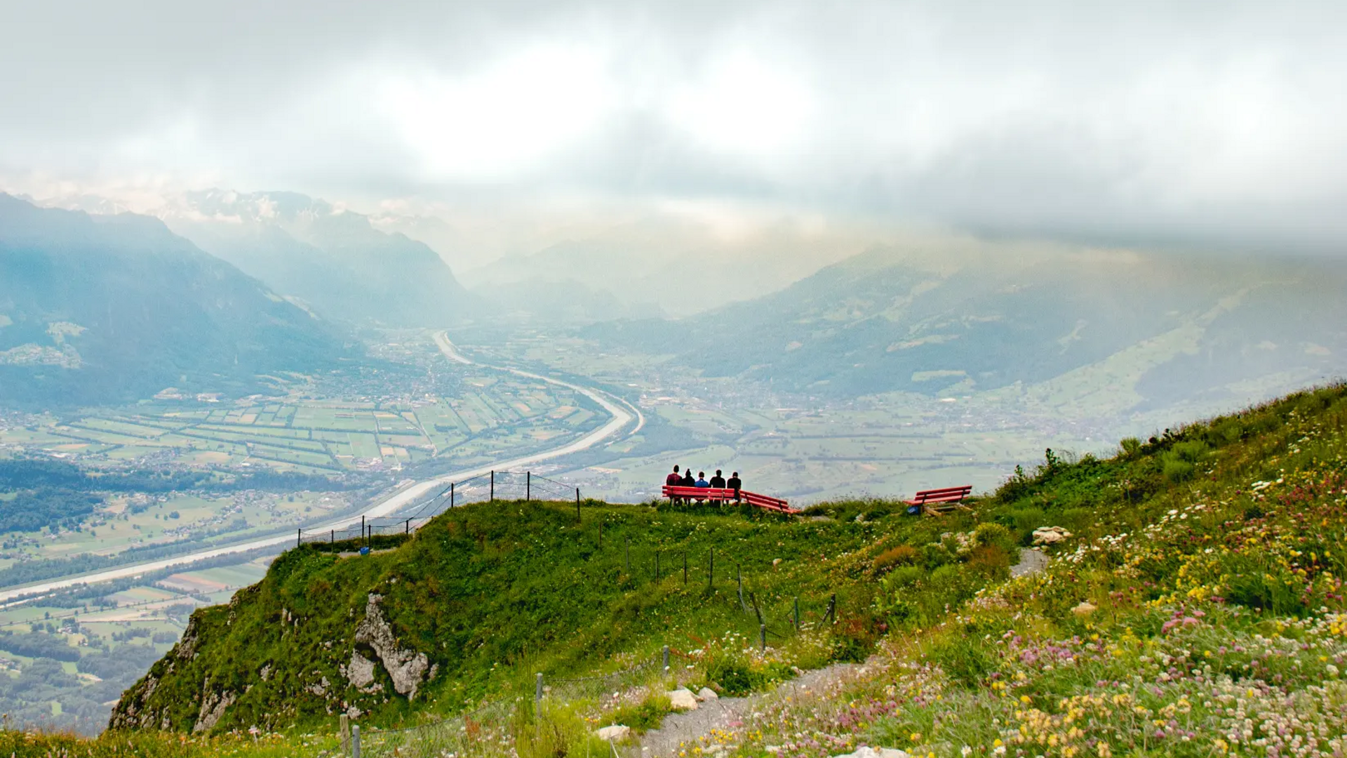People on a bench on green hill overlooking valley and river under cloudy sky