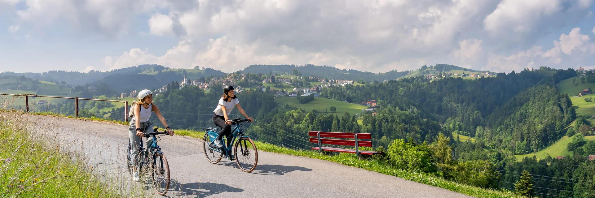 Twee vrouwen fietsen op bergweg met uitzicht op vallei en blauwe lucht