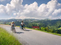 Twee vrouwen fietsen op bergweg met uitzicht op vallei en blauwe lucht
