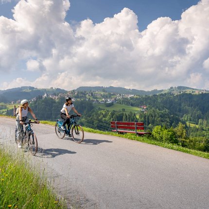 Two women cycling on mountain road with valley view and blue sky